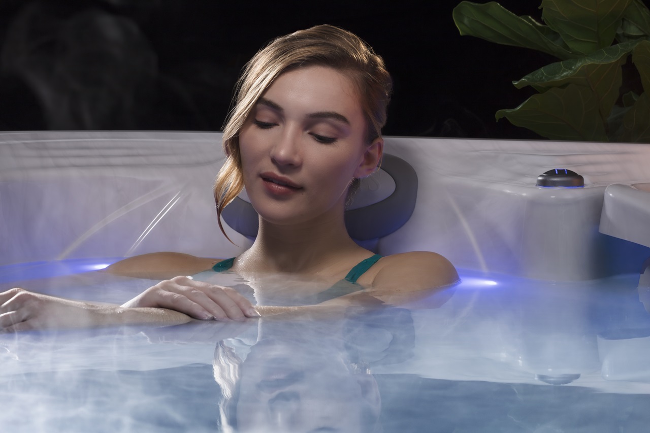 A woman relaxing in a hot tub, leaning back against a headrest with her eyes closed. Soft steam rises from the water, which is illuminated with blue LED lighting.